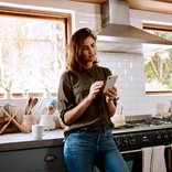 Eine junge Frau steht mit ihrem Smartphone in der Küche.