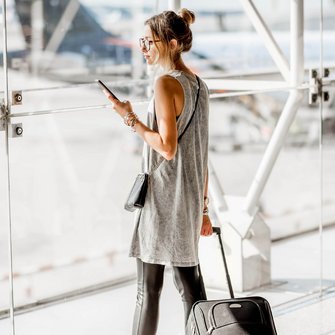 Eine junge Frau steht mit Rollkoffer und Smartphone in der Hand am Flughafen.
